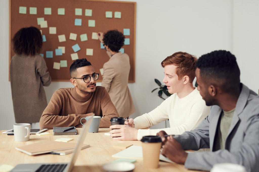 three men sit together at a table in discussion, two women standing in back planning at a corkboard with post it notes